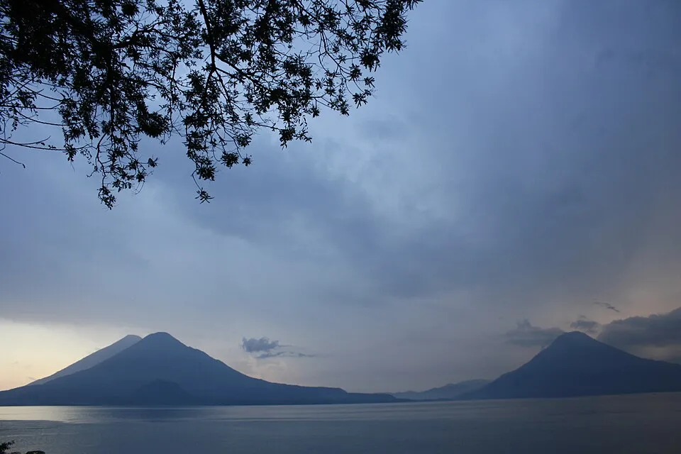 Twin volcano peaks at dusk seen across Lake Atitlan, with tree branches silhouetted in the foreground.