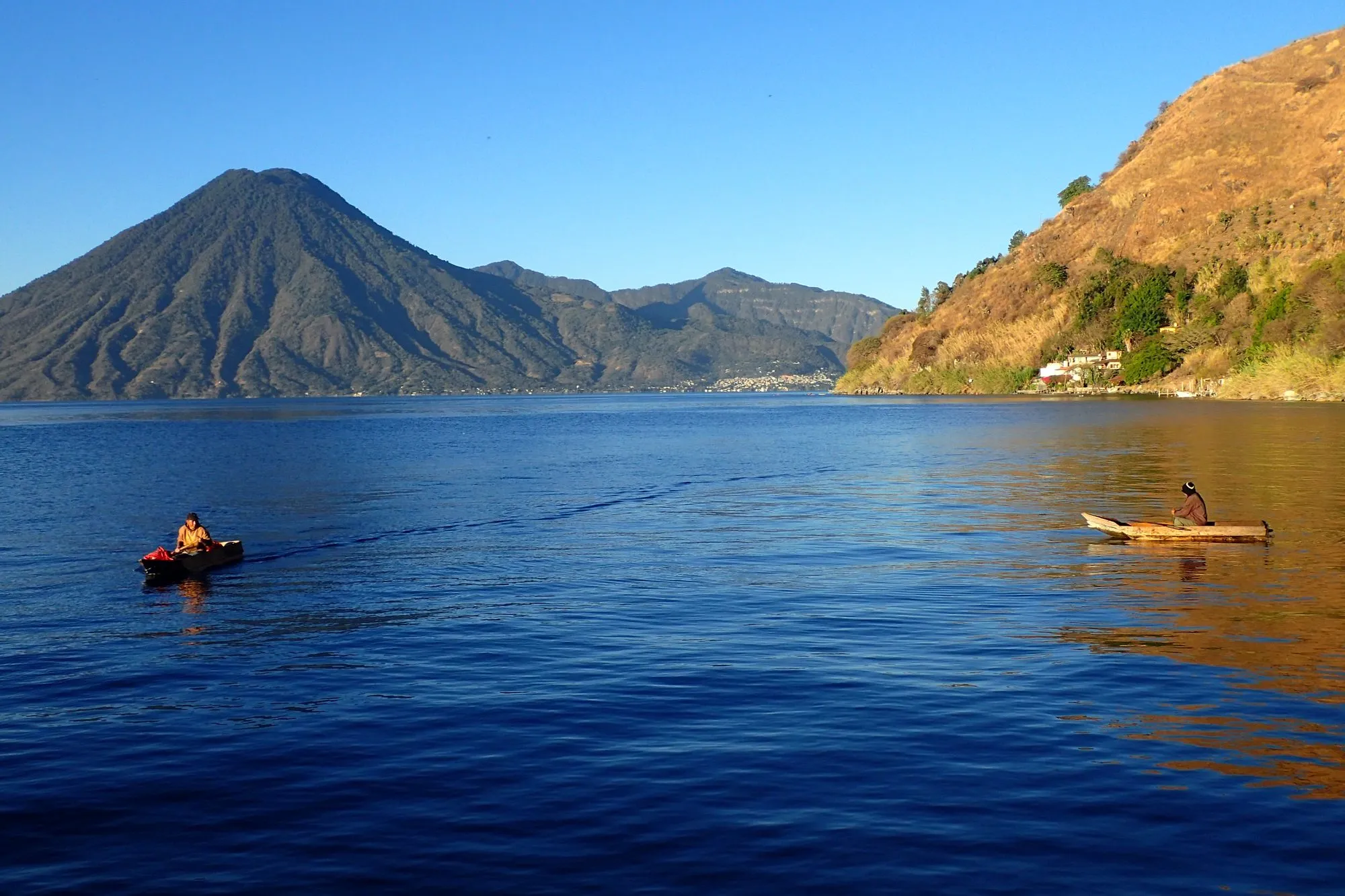 Two small wooden boats with fishermen crossing Lake Atitlan at golden hour near Jaibalito, with a volcano silhouette in the distance.