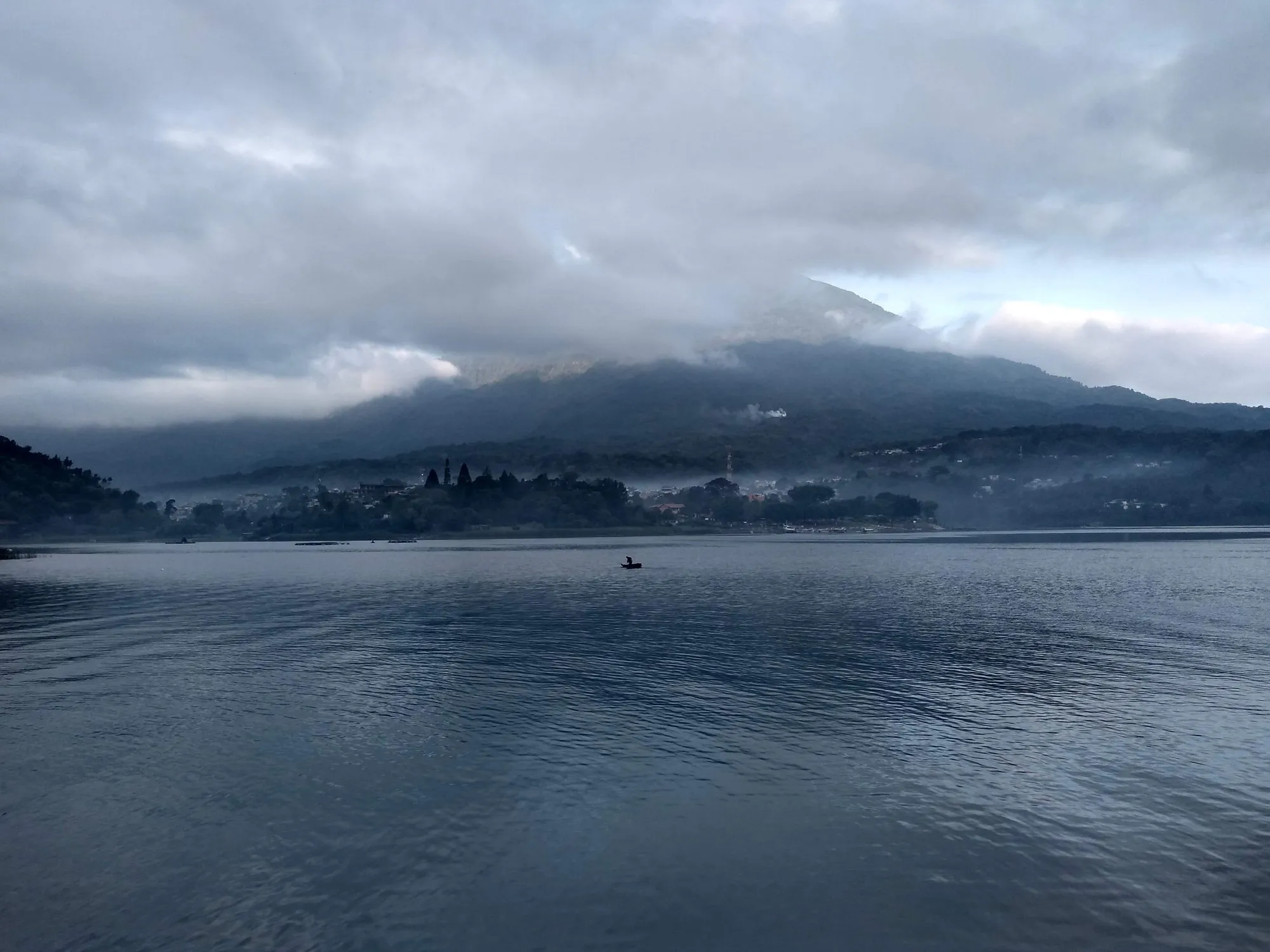 Un pescador solitario en una lancha pequeña en el Lago de Atitlán al amanecer, con un volcán parcialmente cubierto de nubes bajas y un pueblo junto al lago.