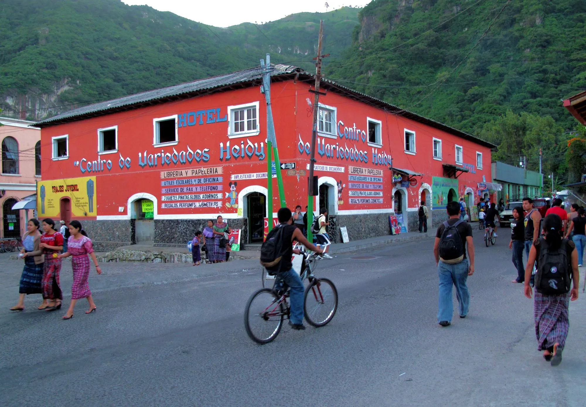 A red two-story building with shops and a hotel sign on a busy Panajachel street, with people walking and cycling past.