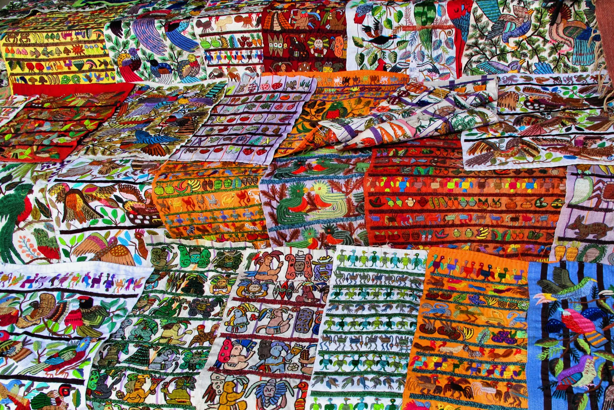 Colorful hand-embroidered textiles with bird and plant motifs laid out for sale, a craft associated with Santiago Atitlan.